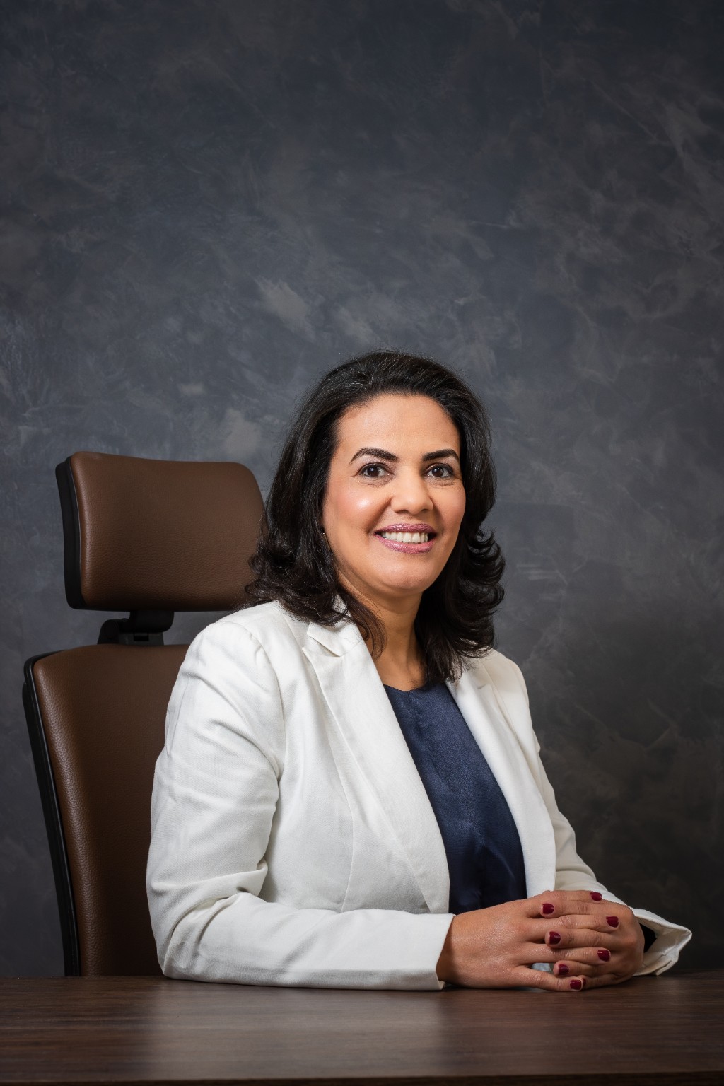 Gilmara Sena, de blazer branco, sentada à mesa e sorrindo